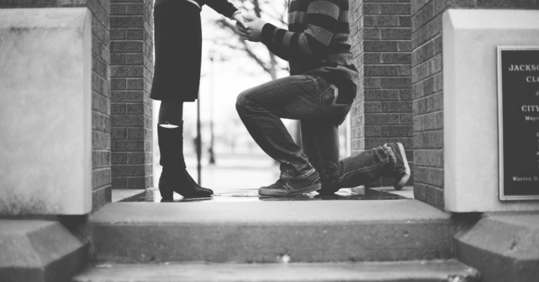 Romantic moment of Man proposing on one knee, holding partner’s hand, outdoor in black and white.